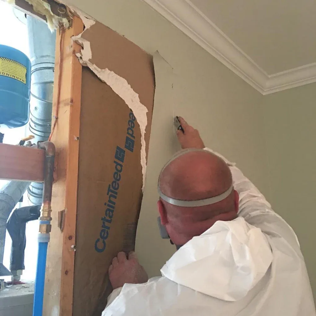 A technician in a protective suit cutting away drywall next to exposed pipes and a pressure tank